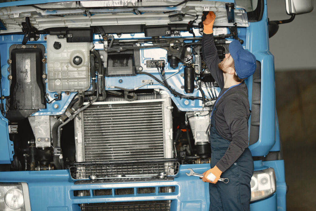 Reduce fleet maintenance costs by performing regular preventive maintenance, shown by a mechanic inspecting and repairing a truck engine to prevent breakdowns and costly repairs.