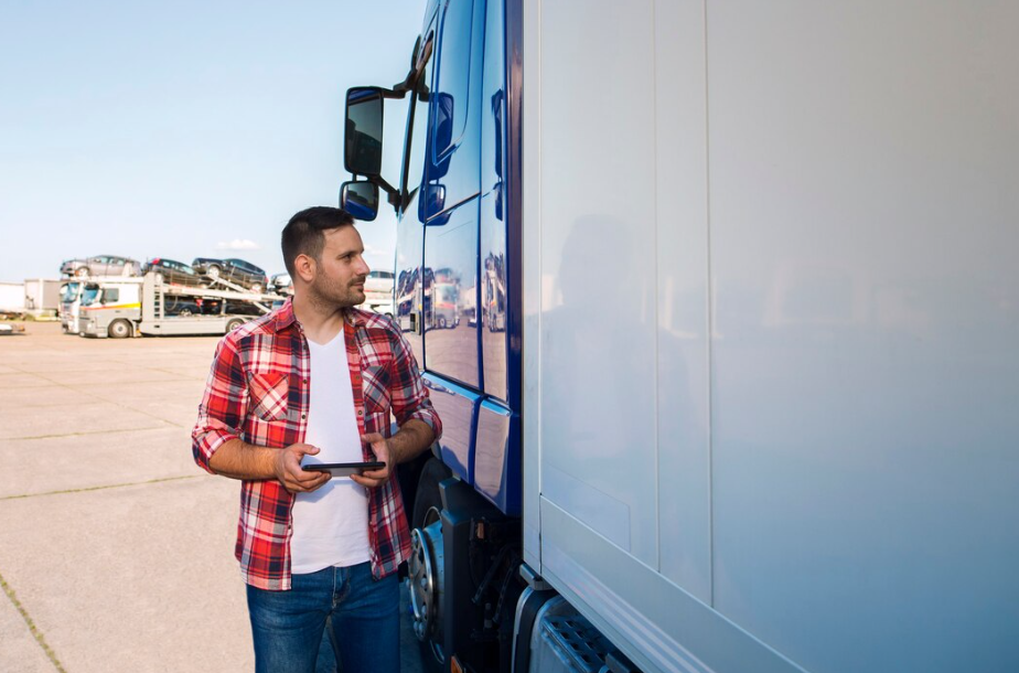 Truck driver standing beside a blue semi in a loading yard, using a tablet to check logs and handling ELD exceptions during trip planning.
