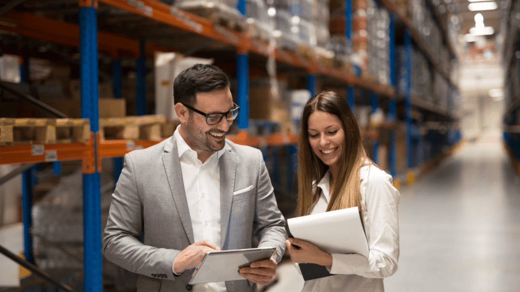 Two professionals reviewing documents and a tablet in a warehouse aisle, representing freight broker coordination, logistics planning, and supply chain communication.